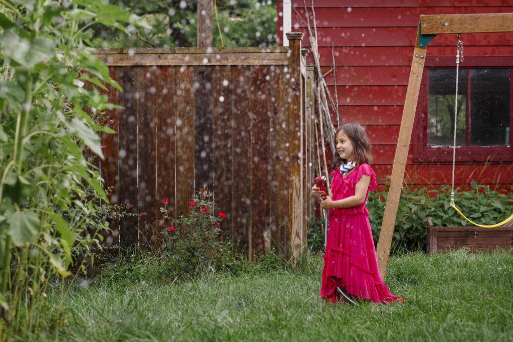 A little girl in long colorful dress plays with hose in backyard