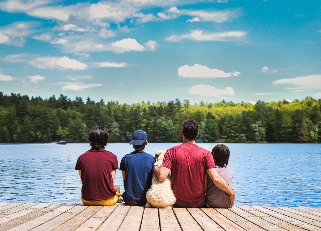 Father, sons and dog sitting on dock on a lake on a sunny summer day.