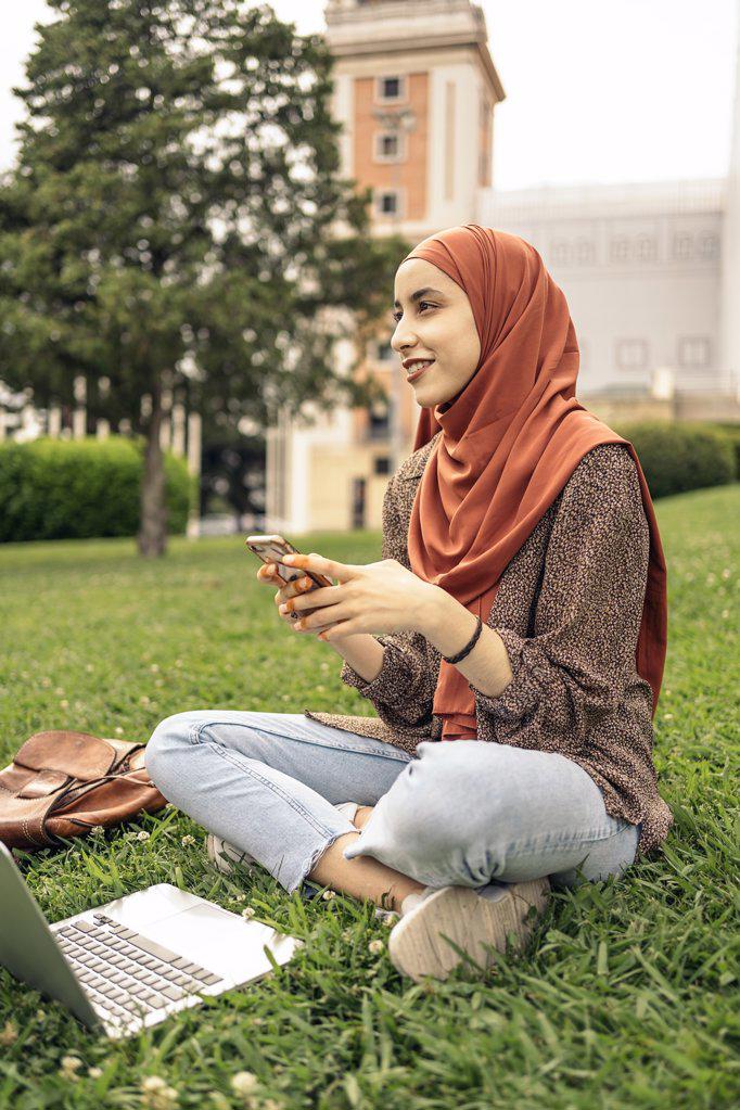 Muslim woman siting on the grass using a computer and phone