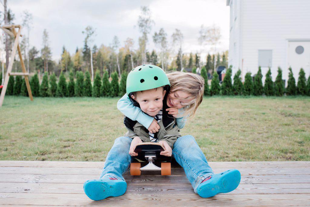 sister hugging her brother whilst playing on a skateboard outside