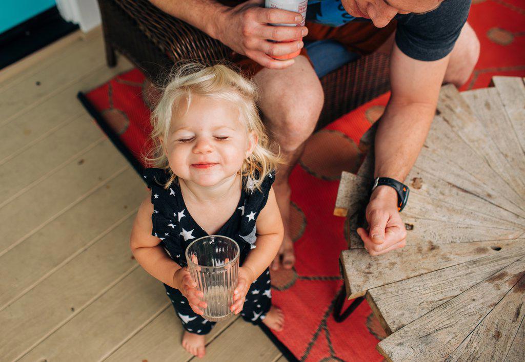 Blonde toddler girl drinks happily from cup while dad watches