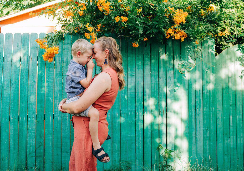 Mother snuggles little boy while standing in front of green fence
