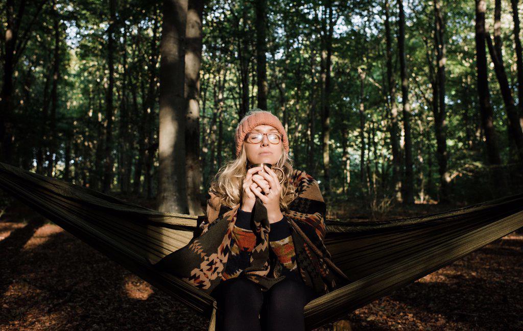 woman sat on a hammock in the forest enjoying a hot drink in autumn