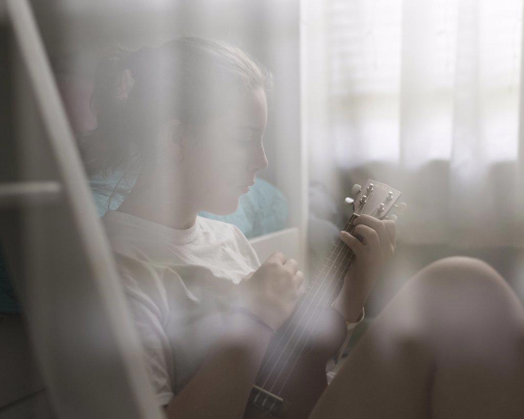 Teen girl with ponytail plays ukulele sitting by white bunkbed