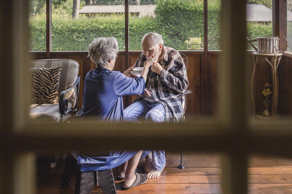 Female caretaker feeds feeble elderly man on screened porch