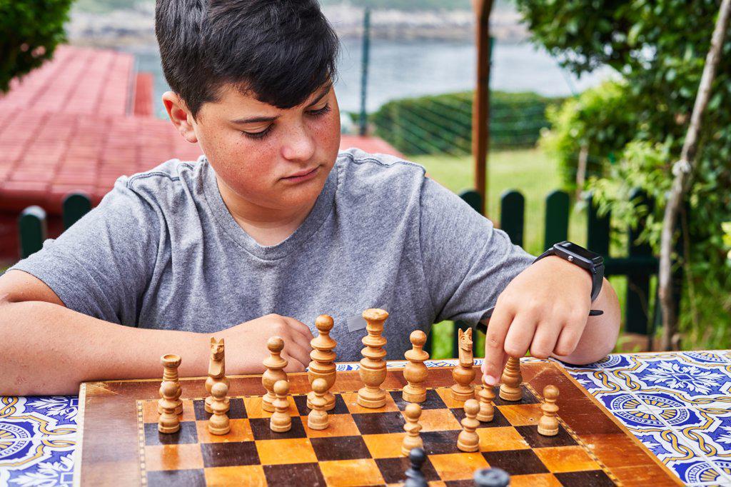 Young pensive boy sitting behind chess board moving one of the pieces.