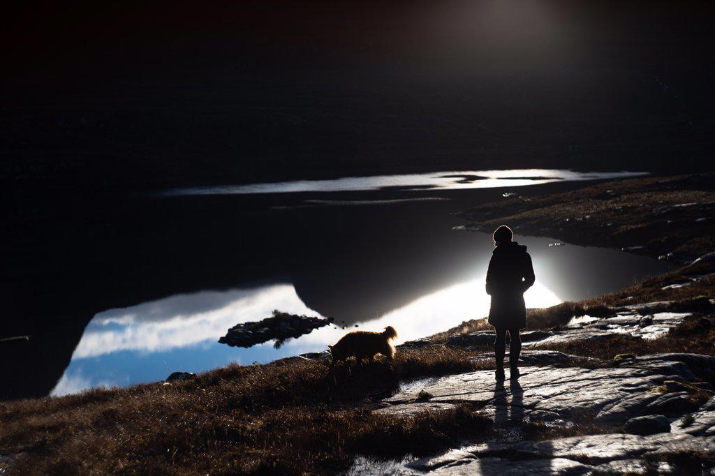 Silhouette of woman and dog against mountain reflection in lake