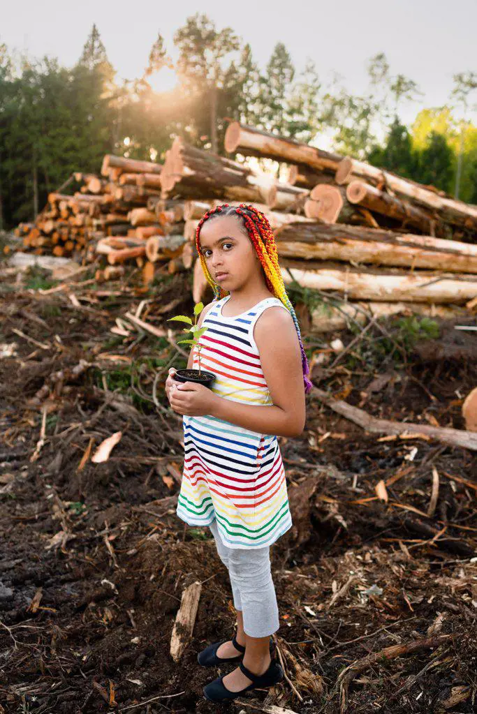 Girl with rainbow braids holds sapling on logging site