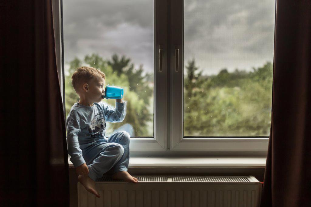 Blonde boy in blue pajamas drinking milk and sitting on windowsill