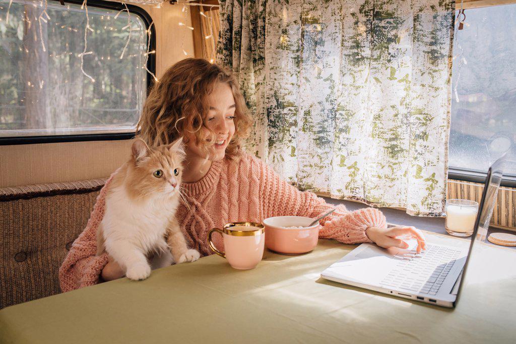 Woman and ginger cat in trailer are eating breakfast at laptop.