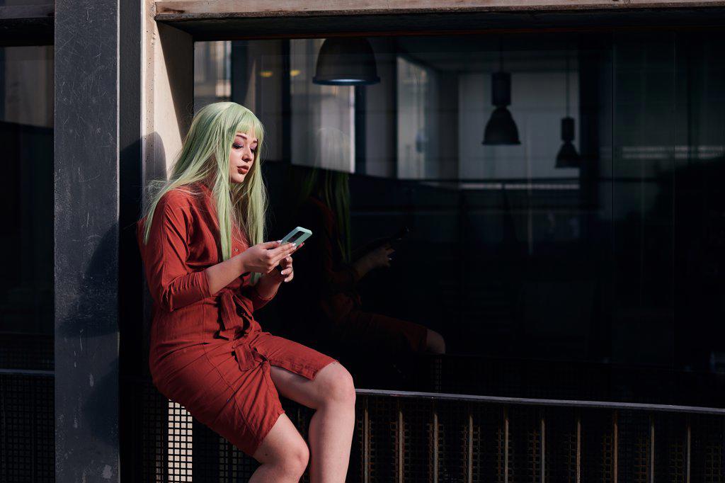 Person in a green wig and red dress looks at her phone on the street