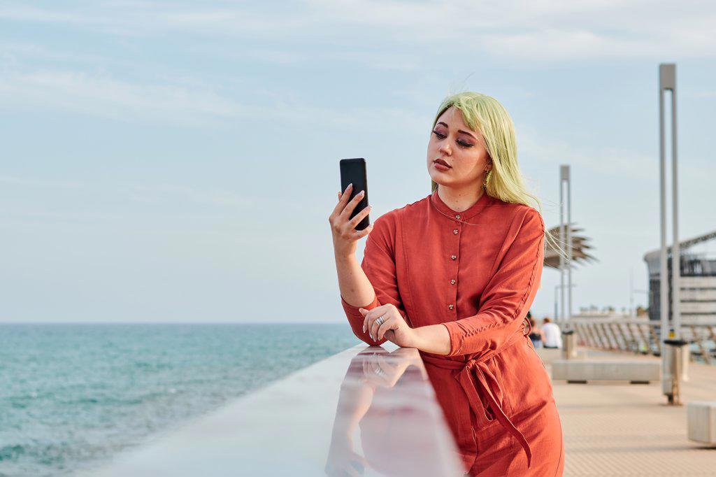 Non-binary person looks at her smartphone with the sea in the back
