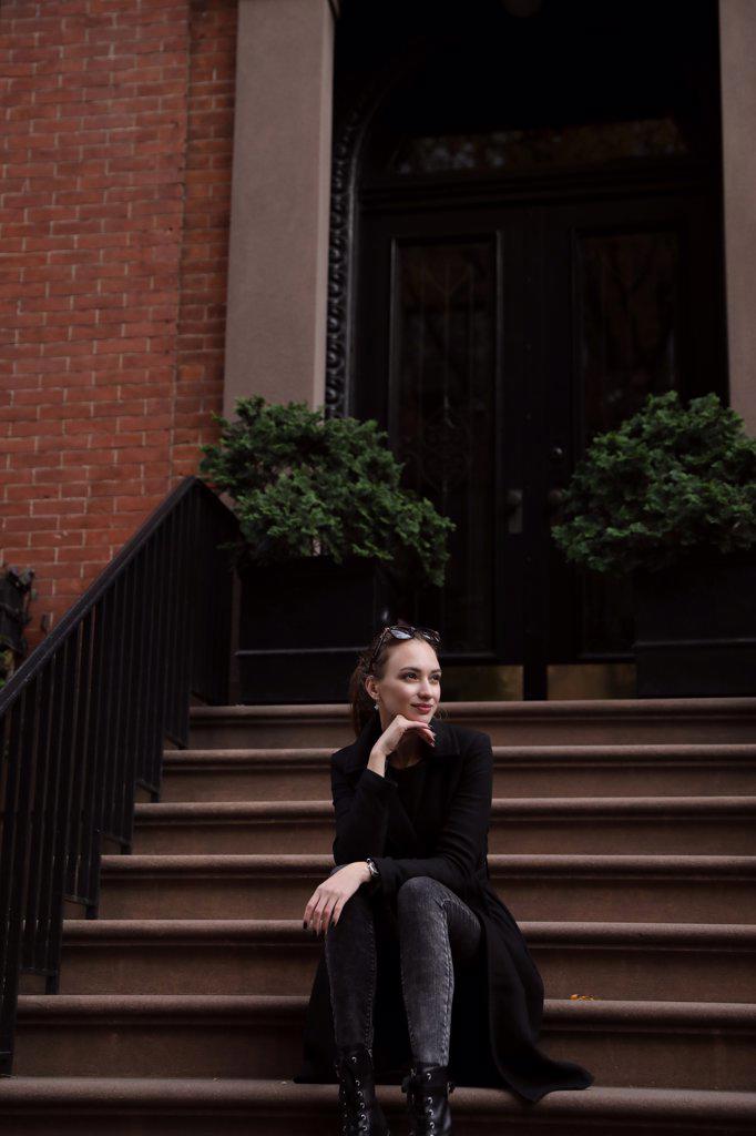 Young woman in black coat sits on the steps of the house
