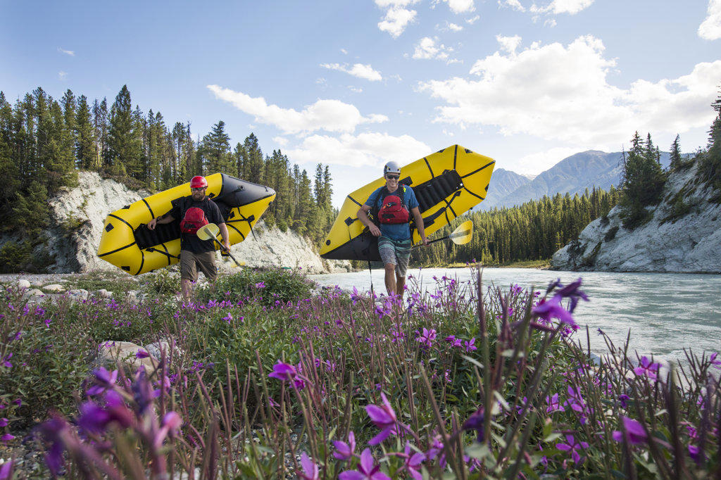 Paddlers carry yellow packrafts beside scenic river, flowers