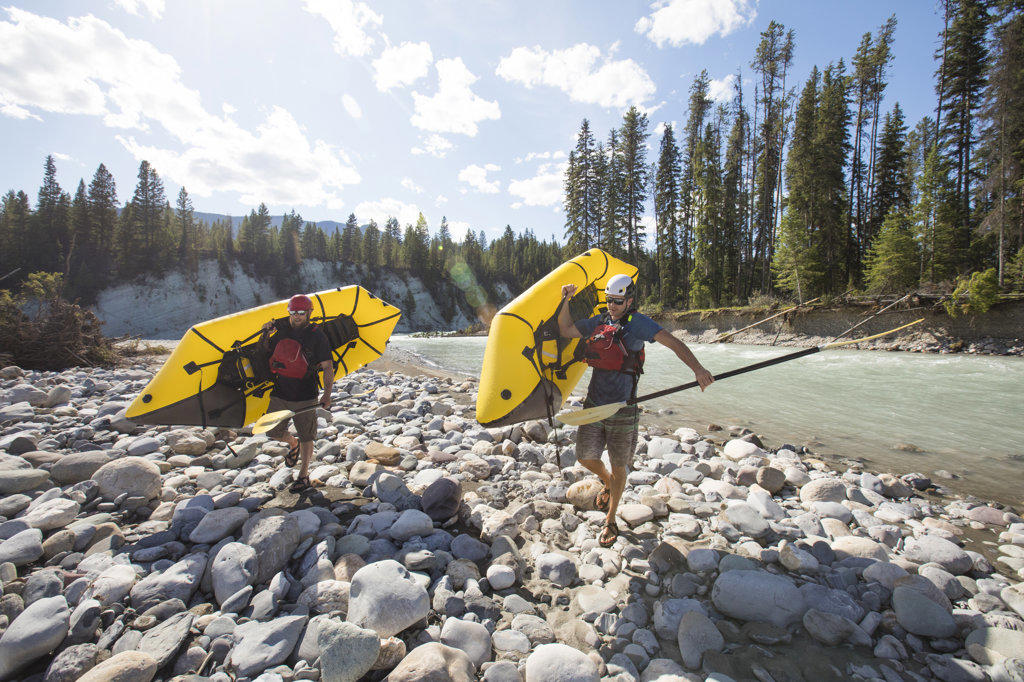 Two men carry inflatable rafts on a river adventure.