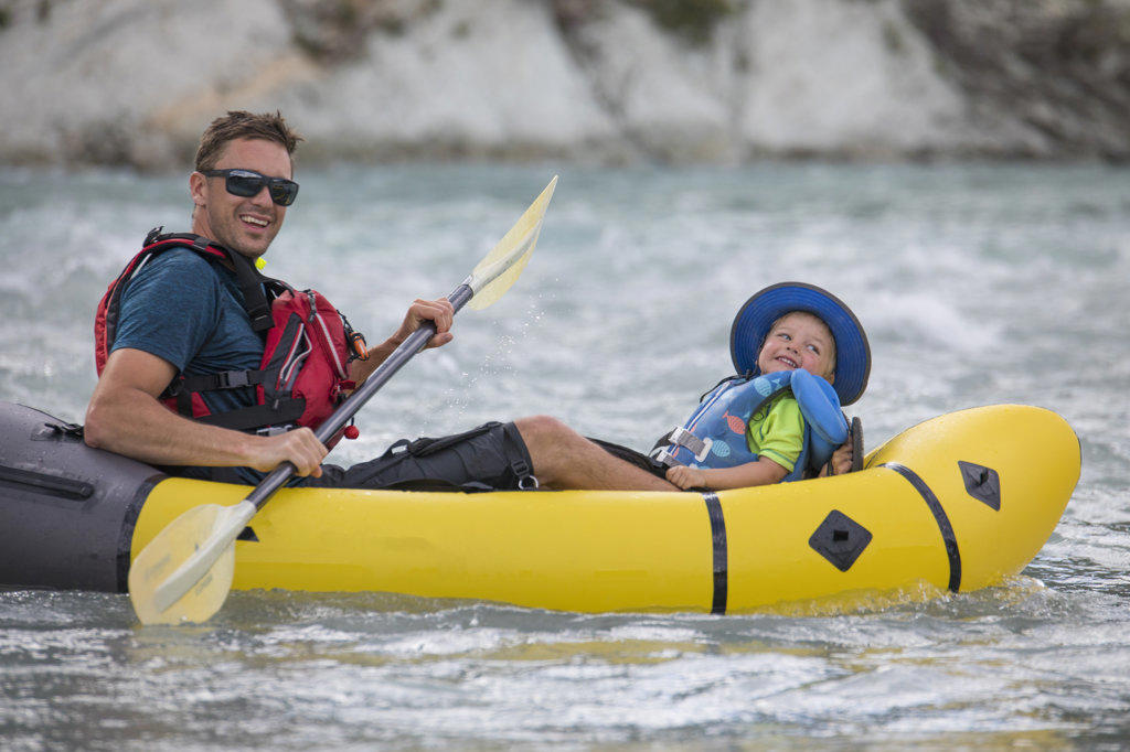 Father and son paddle river on a packraft, wearing lifejackets.