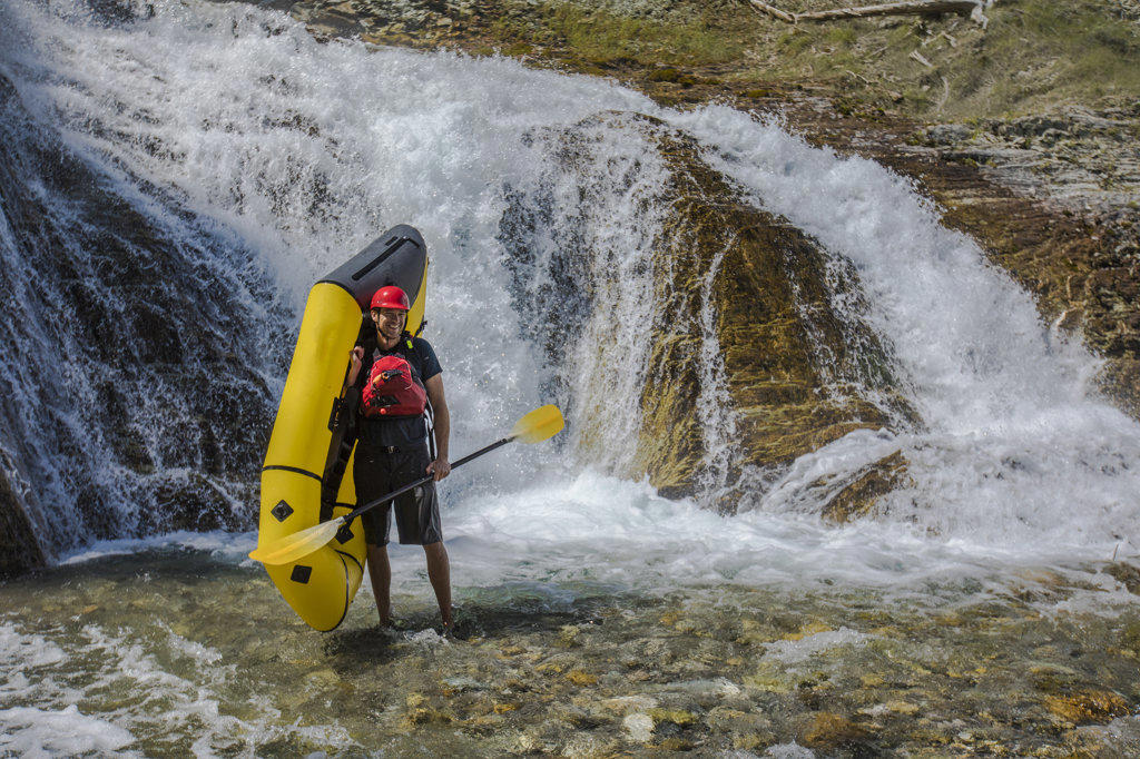 Man holds packraft in river next to waterfall.