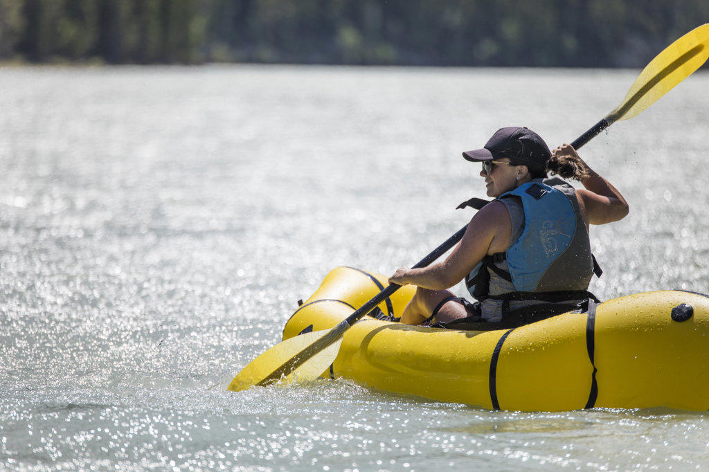 Side view of fit active woman paddling yellow packraft on river.