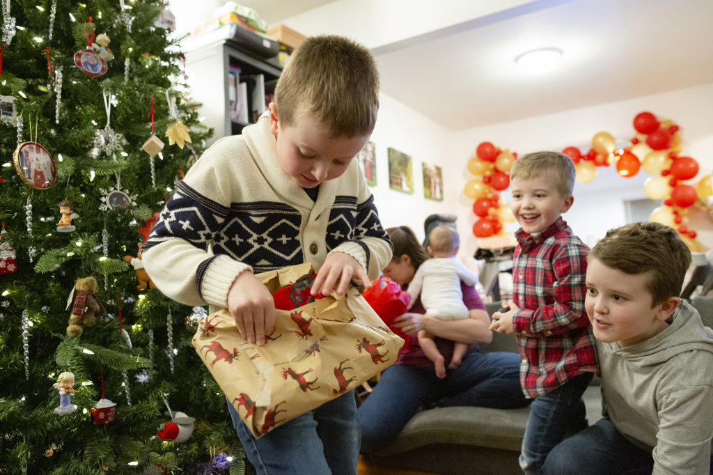 Boy Opens Christmas Present in Front of Tree While Brothers Look On