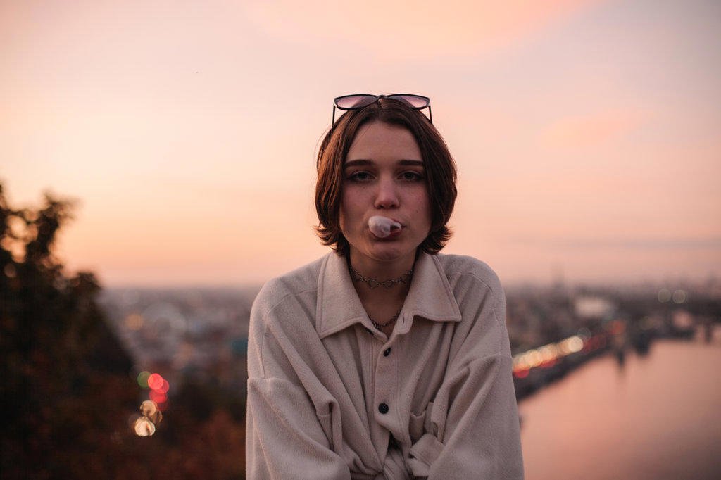 Young woman blowing bubble gum against city at sunset
