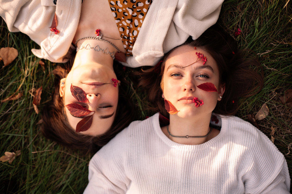 Two young women with red leaves on faces lying on grass in autumn