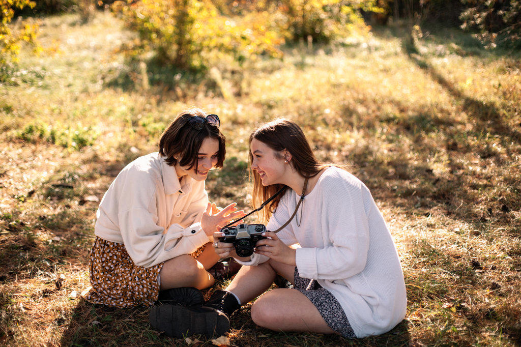 Happy female friends with camera sitting on grass in park in autumn