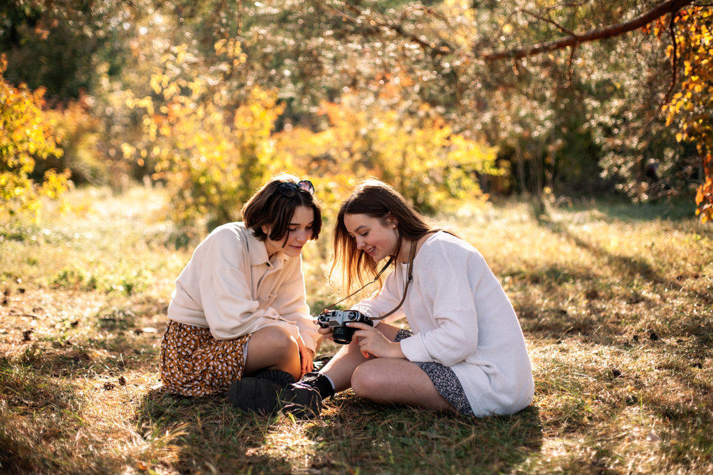 Two teenage girls with camera sitting on grass in park in autumn