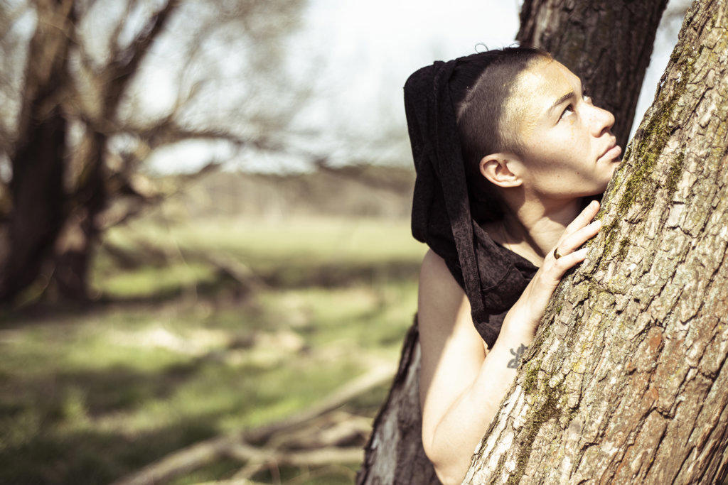 non-binary person with hood stands in trees looking up outdoors