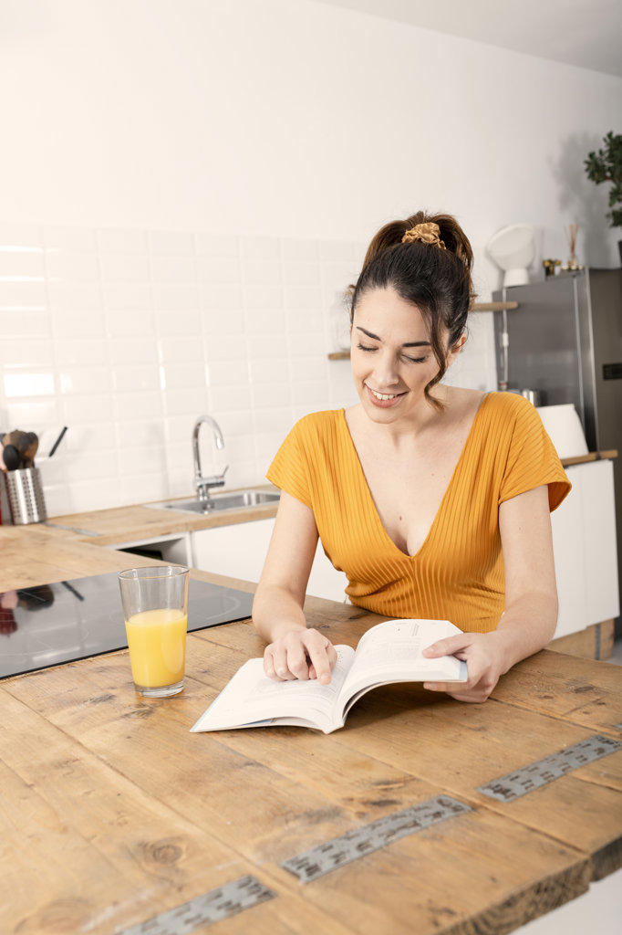 Beautiful young woman reading book in kitchen near orange juice