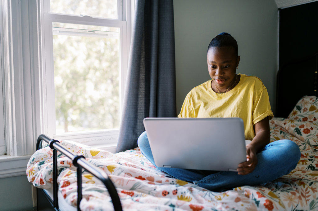 young laughing black teen sitting on bed with computer