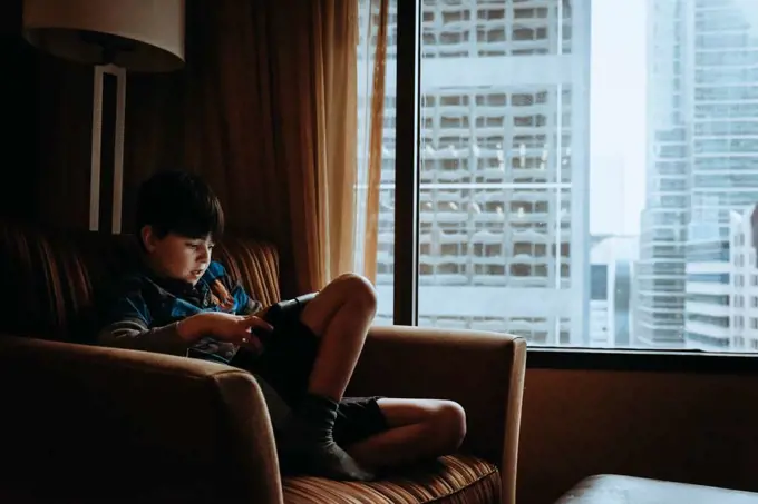 Boy using tablet in chair beside window with tall buildings outside.