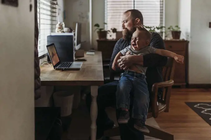 Dad working from home with one year old boy crying in his lap