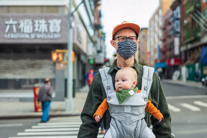 Father wearing face mask carrying daughter in baby carrier while crossing street in city during pandemic