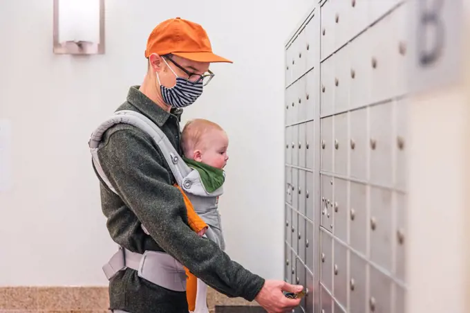Father wearing face mask opening locker in room while carrying baby girl during COVID-19 crisis