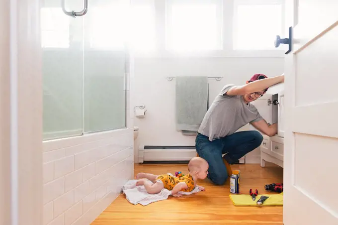 Man fixing sink while daughter lying on hardwood floor in kitchen