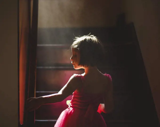 A graceful child stands in hazy light on dark staircase in fancy dress