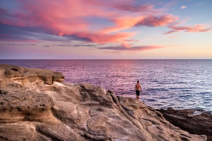 Fit male stands at edge of cliff during pink hawaii sunset