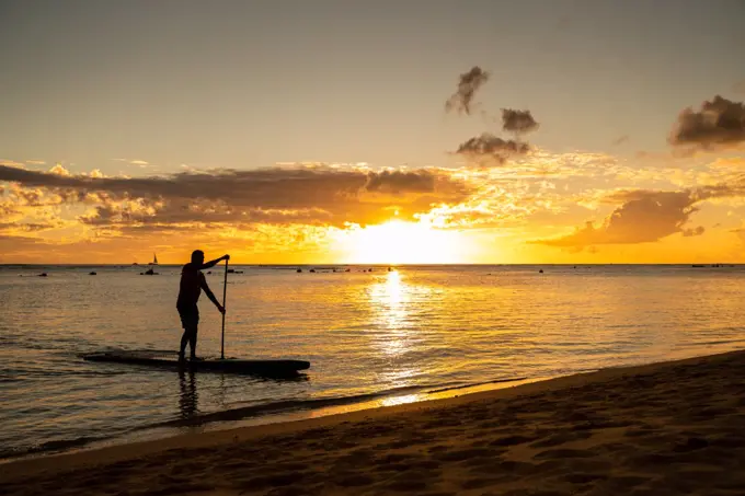 Male stand up paddle boarding drifts to the sand at sunset