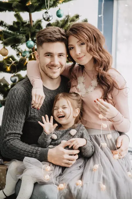 Young family with a daughter in festive outfits with a garland near the Christmas tree on New Years Eve