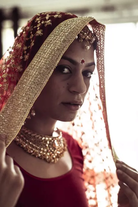 Young Indian woman pulls on red wedding veil in window light