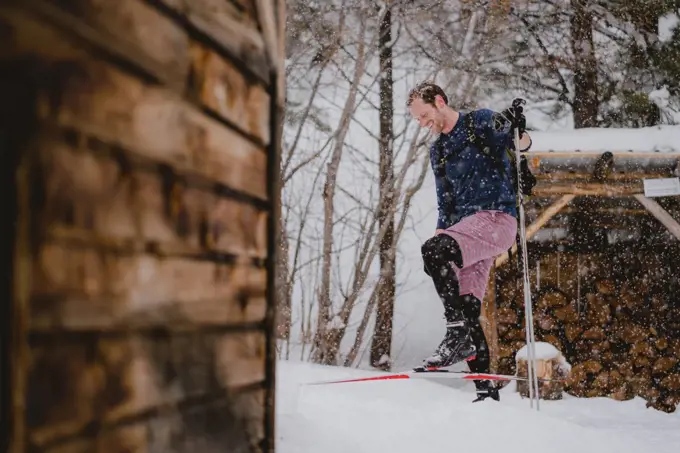 A man laughs in the snow while XC skiing outside of a rustic cabin