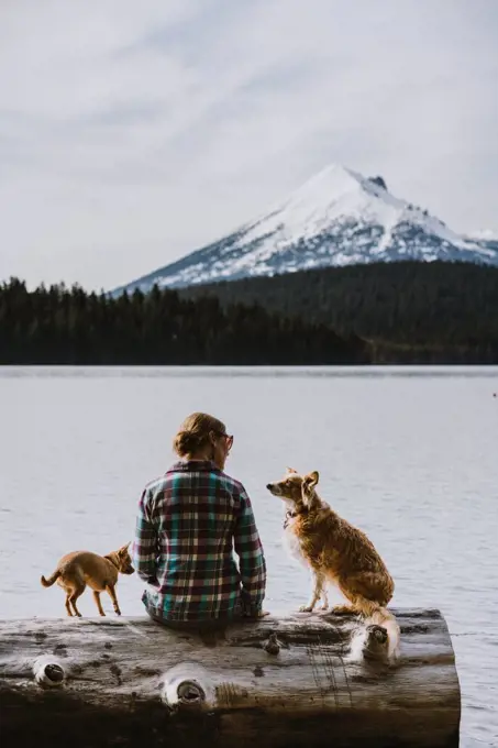A woman sits and enjoys view of lake and mountains with her two dogs