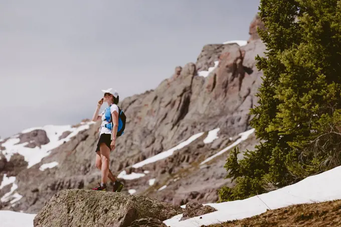 Woman trail runner sips water in front of pine trees snow and rocks