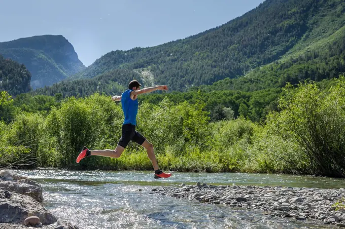 Male trail runner leaps over rocky river on sunny day in the mountains