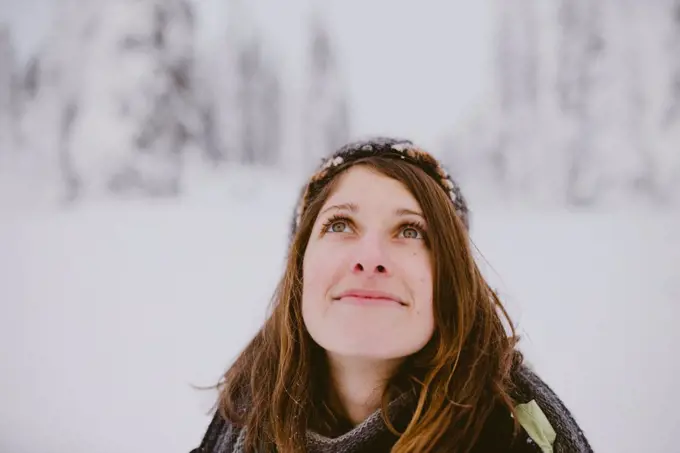 Young woman with brown hair and eyes looks up at snow covered trees