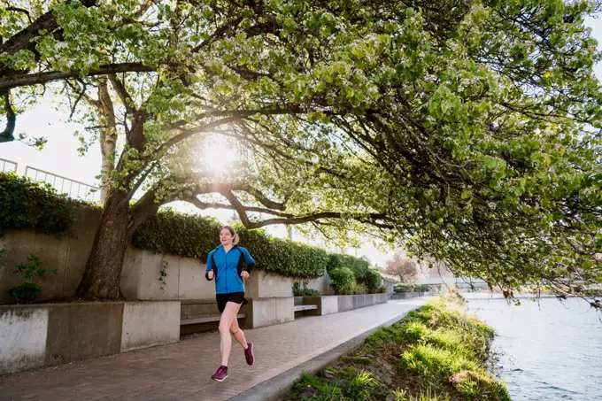 Young woman runs on paved path through a tree tunnel next to river