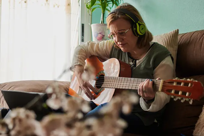 Middle-aged woman with a headphones plays guitar on the sofa at home