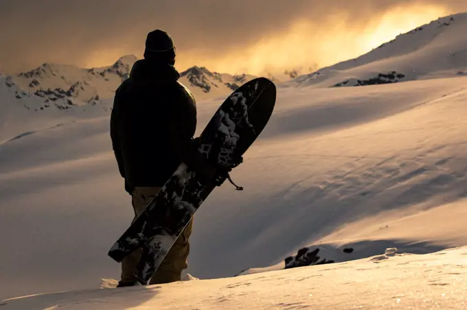 Rear view of man with snowboard standing on snowcapped mountain during sunset