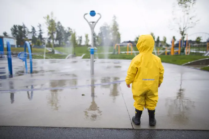 Toddler in rain suit and boots looks at the water park on a wet day