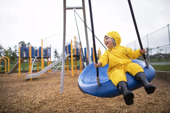 Happy toddler boy rides on swing at the park on a wet day